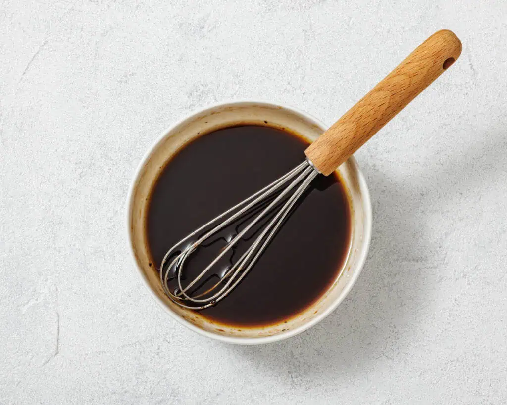 A metal whisk rests in a white bowl filled with a dark liquid, possibly a sauce or marinade, on a light textured surface.