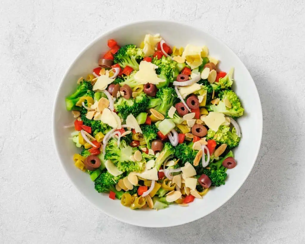 A bowl of broccoli salad with sliced olives, red bell peppers, onions, shaved cheese, and slivered almonds on a light surface.