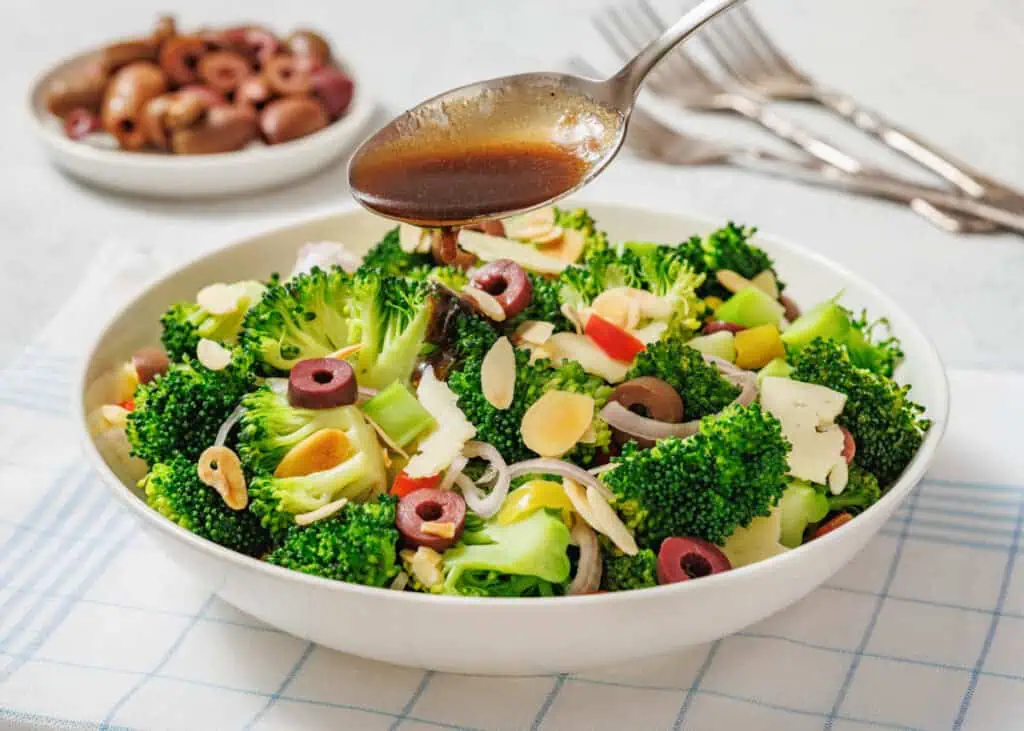 A bowl of broccoli salad with olives, sliced almonds, and vegetables is being drizzled with dressing from a spoon. A plate of olives and forks are in the background.