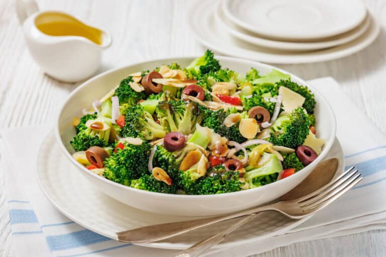 A bowl of Italian broccoli salad with sliced olives, almonds, onions, and red peppers, served on a white plate with a fork and napkin beside it.