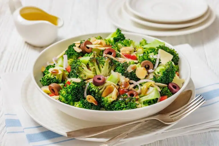 A bowl of Italian broccoli salad with sliced olives, almonds, onions, and red peppers, served on a white plate with a fork and napkin beside it.