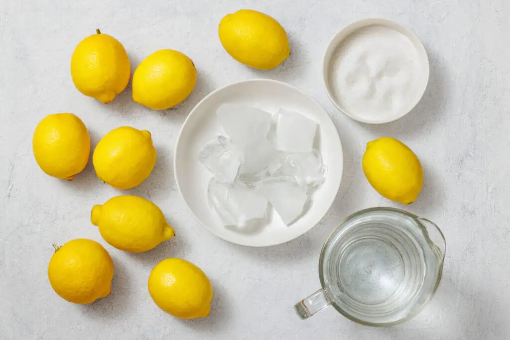 Flat lay of fresh lemons, a bowl of sugar, ice cubes on a plate, and a glass pitcher of water, arranged on a light-colored surface, showcasing the ingredients for homemade lemonade.
