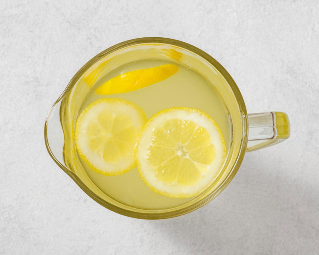 Top-down view of a clear pitcher with lemonade and lemon slices on a light gray surface.