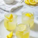 Three glasses of lemonade with ice and lemon slices on a white plate, with a bowl of sugar and sliced lemons in the background.