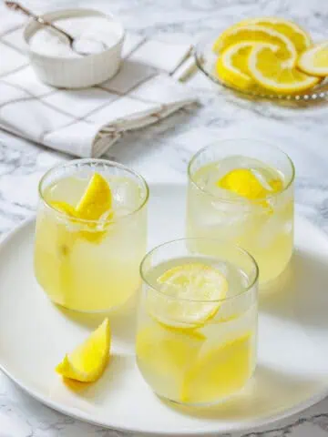 Three glasses of lemonade with ice and lemon slices on a white plate, with a bowl of sugar and sliced lemons in the background.