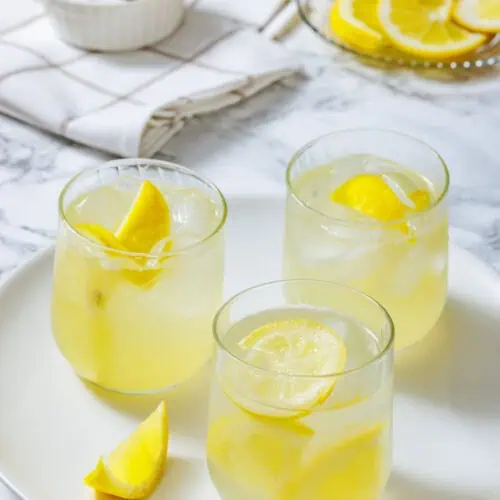 Three glasses of lemonade with ice and lemon slices on a white plate, with a bowl of sugar and sliced lemons in the background.