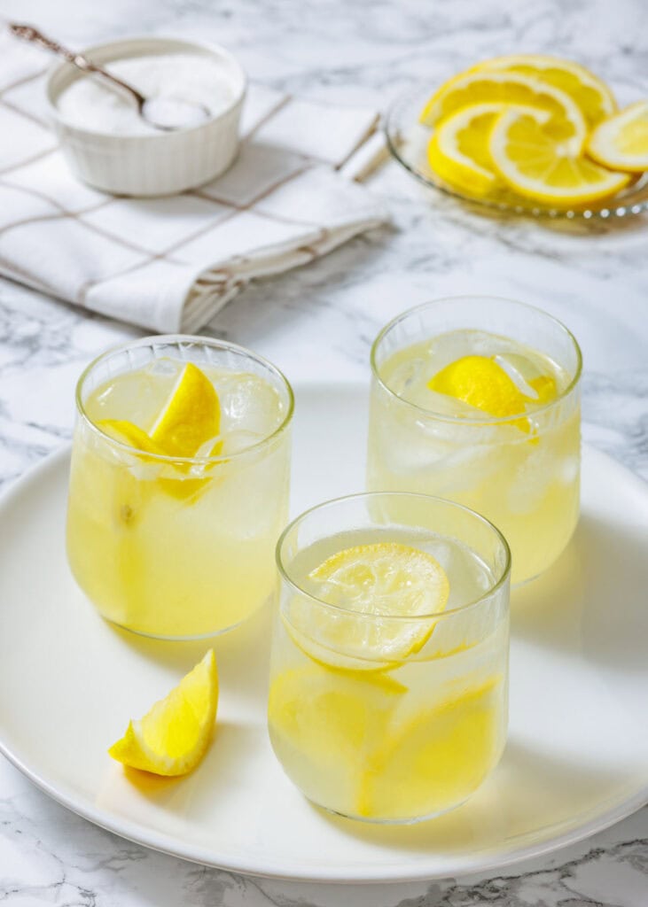 Three glasses of lemonade with ice and lemon slices on a white plate, with a bowl of sugar and sliced lemons in the background.