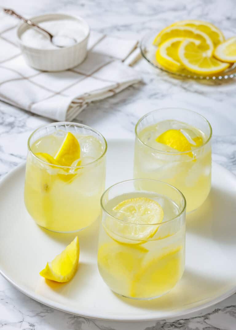 Three glasses of lemonade with ice and lemon slices on a white plate, with a bowl of sugar and sliced lemons in the background.