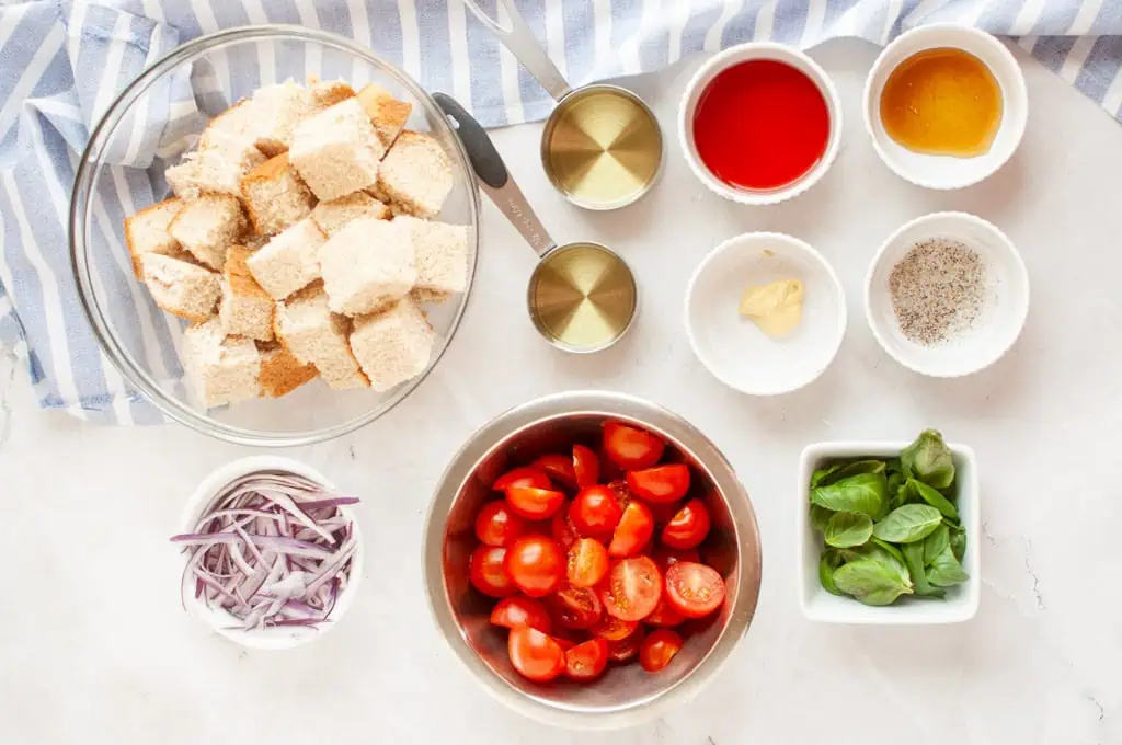 Overhead shot of panzanella salad ingredients, including bread cubes, tomatoes, red onion, basil, and dressing components, arranged on a light surface.