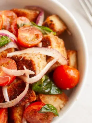 A white bowl filled with panzanella salad, featuring toasted bread cubes, cherry tomatoes, red onion, and basil leaves, is shown with a fork.