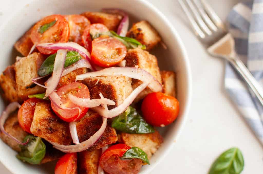 A white bowl filled with panzanella salad, featuring toasted bread cubes, cherry tomatoes, red onion, and basil leaves, is shown with a fork.