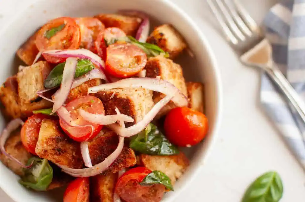 A white bowl filled with panzanella salad, featuring toasted bread cubes, cherry tomatoes, red onion, and basil leaves, is shown with a fork.