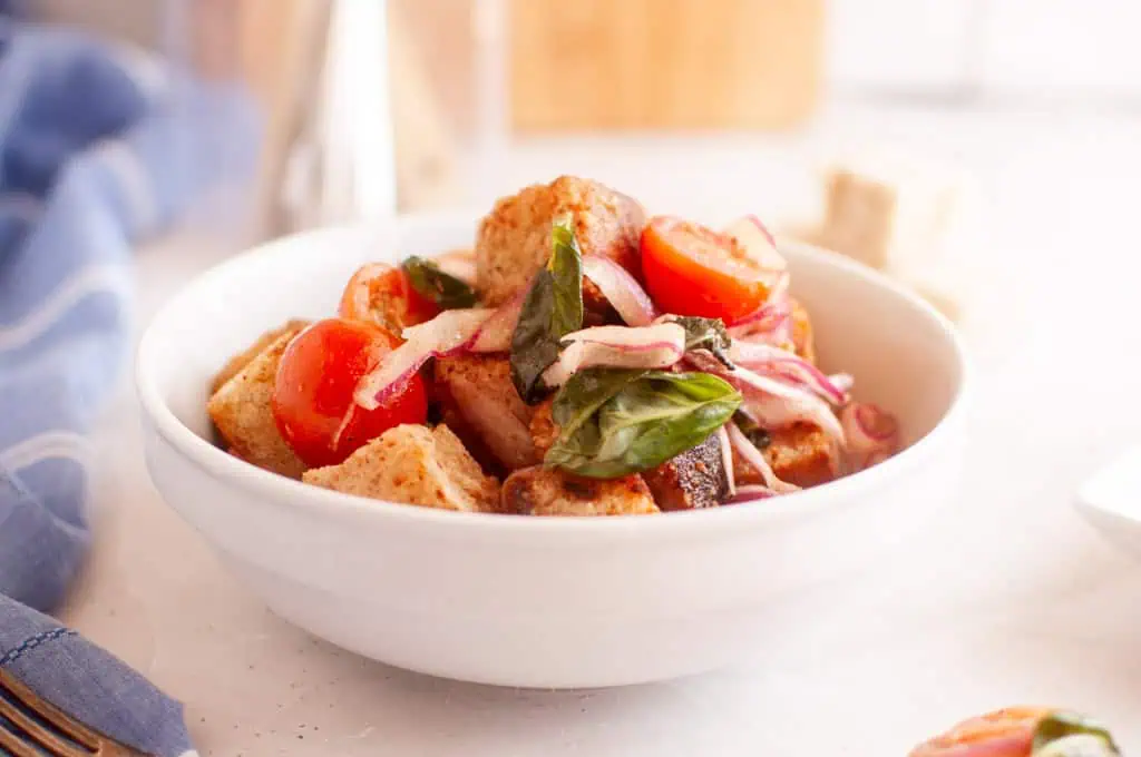 A white bowl filled with panzanella salad, featuring toasted bread cubes, cherry tomatoes, red onion, and basil leaves.