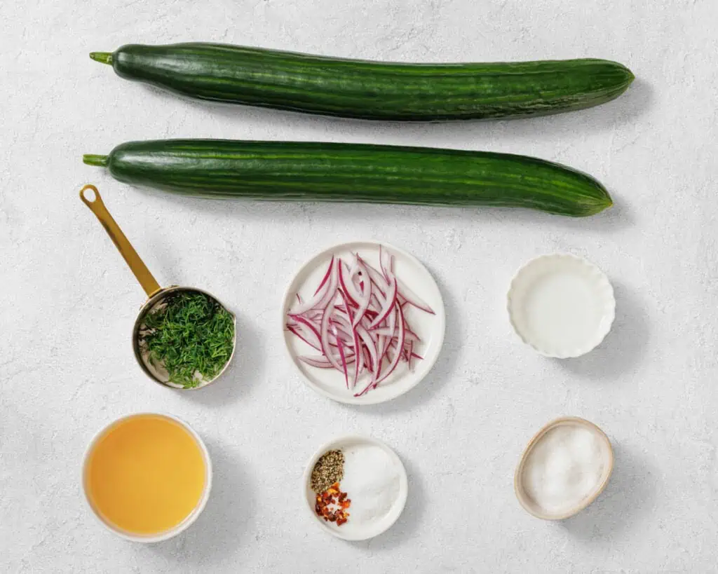 Two whole cucumbers, a plate of sliced red onions, chopped herbs, white powder, seasonings, sugar, vinegar, and an empty white bowl arranged on a light surface.