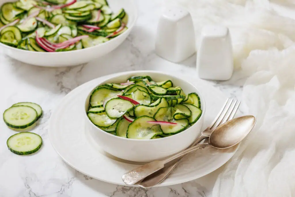 A bowl of Quick Marinated Cucumbers with red onions in a creamy dressing, served on a white plate with a fork and spoon, next to a salt and pepper shaker.
