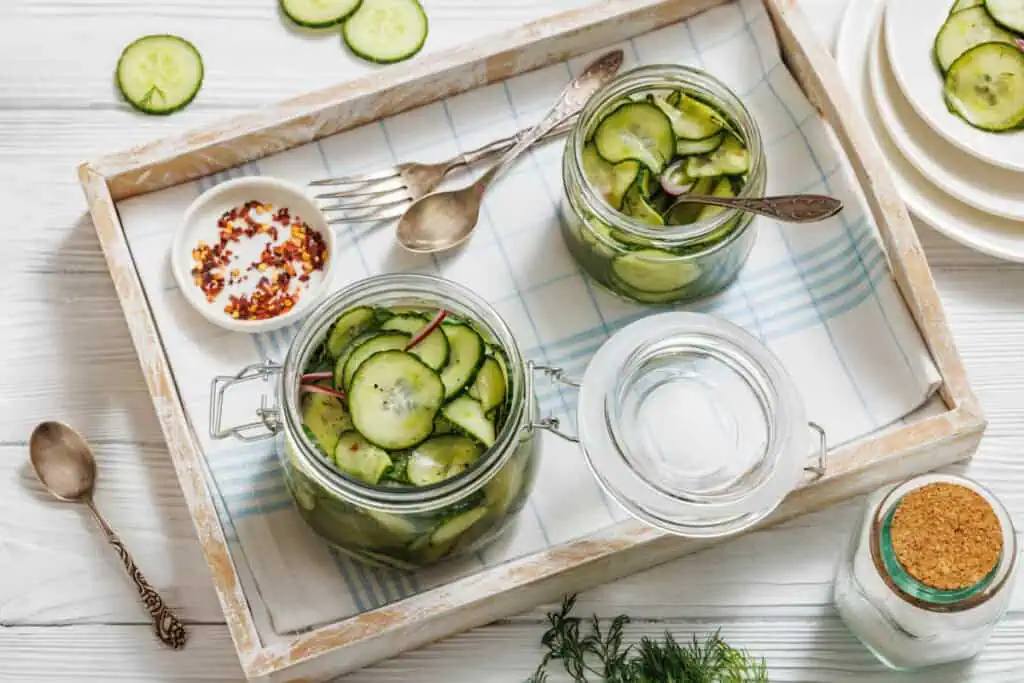 Two jars of Quick Marinated Cucumbers sit on a tray with a bowl of red pepper flakes, spoons, forks, and slices of fresh cucumber on a checkered cloth.