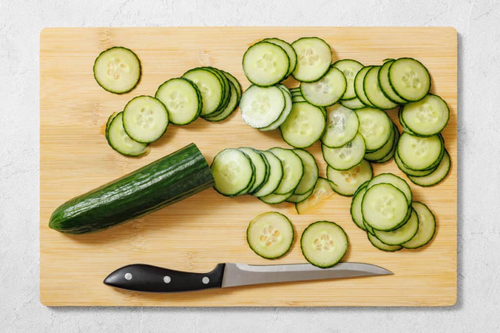 A partially sliced cucumber, cucumber slices, and a knife are arranged on a wooden cutting board.