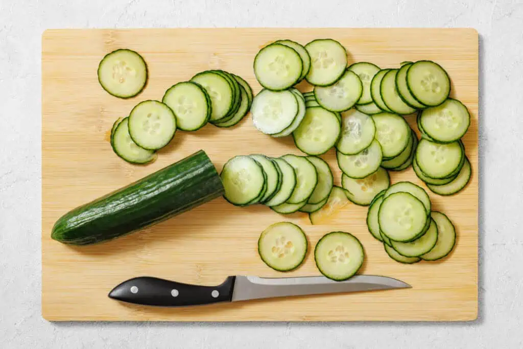A partially sliced cucumber, cucumber slices, and a knife are arranged on a wooden cutting board.