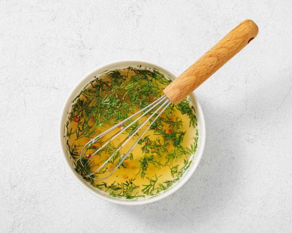 A bowl of liquid with chopped herbs and a whisk with a wooden handle on a light surface.