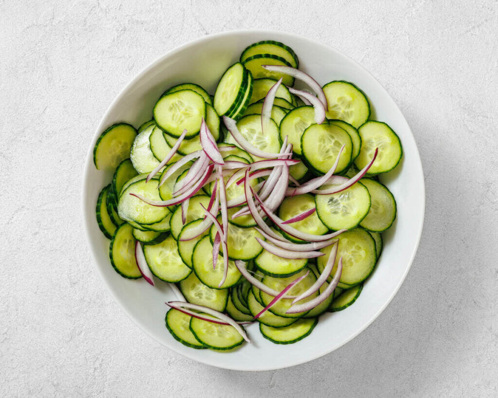 A white bowl filled with sliced cucumbers and thinly sliced red onions on a light gray surface.