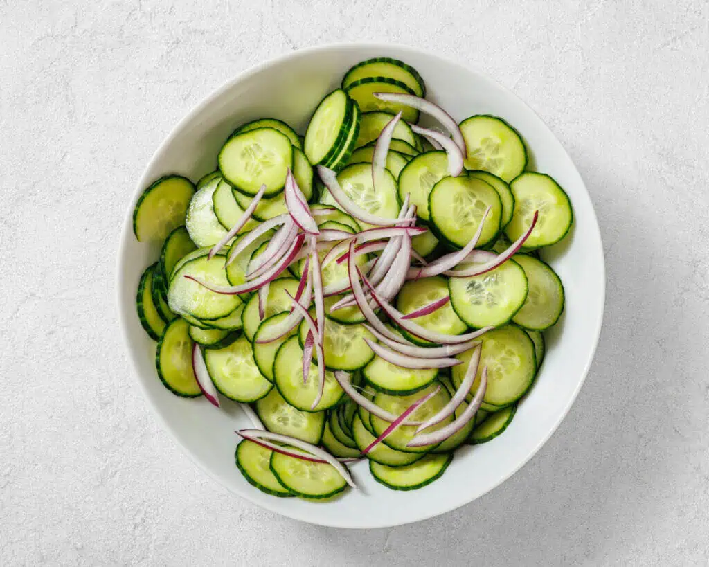A white bowl filled with sliced cucumbers and thinly sliced red onions on a light gray surface.