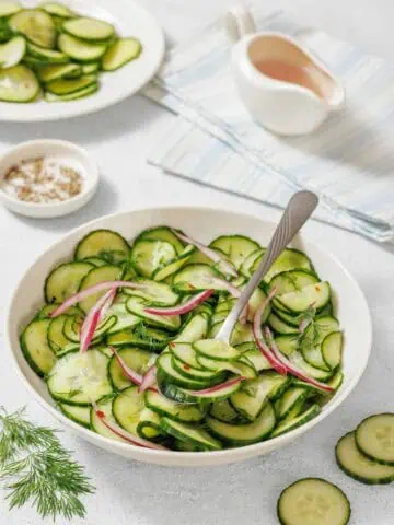 A bowl of Quick Marinated Cucumbers with red onion and dill, a serving spoon, and plates with sliced cucumbers on a light surface. A small dish of salt and dressing are also nearby.