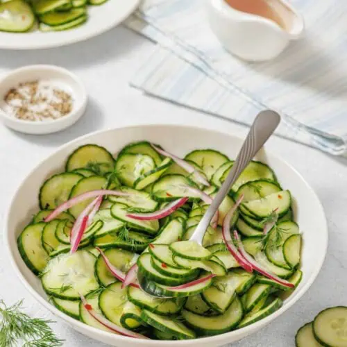 A bowl of Quick Marinated Cucumbers with red onion and dill, a serving spoon, and plates with sliced cucumbers on a light surface. A small dish of salt and dressing are also nearby.