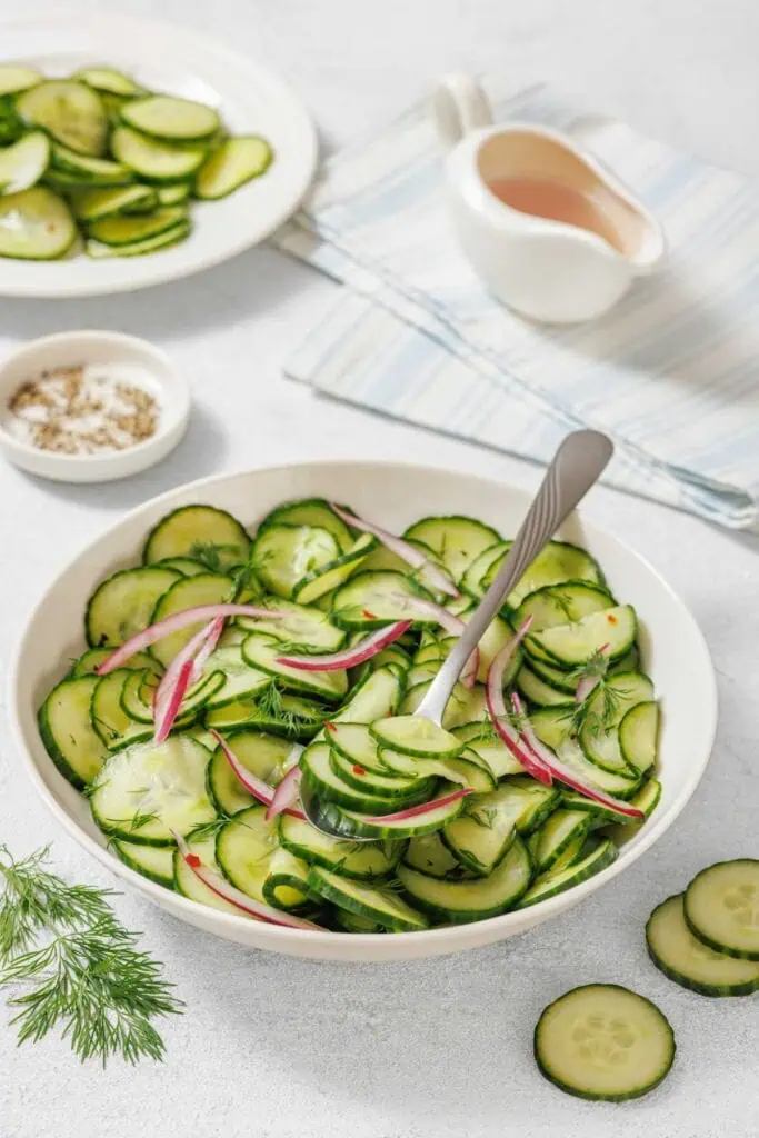 A bowl of Quick Marinated Cucumbers with red onion and dill, a serving spoon, and plates with sliced cucumbers on a light surface. A small dish of salt and dressing are also nearby.