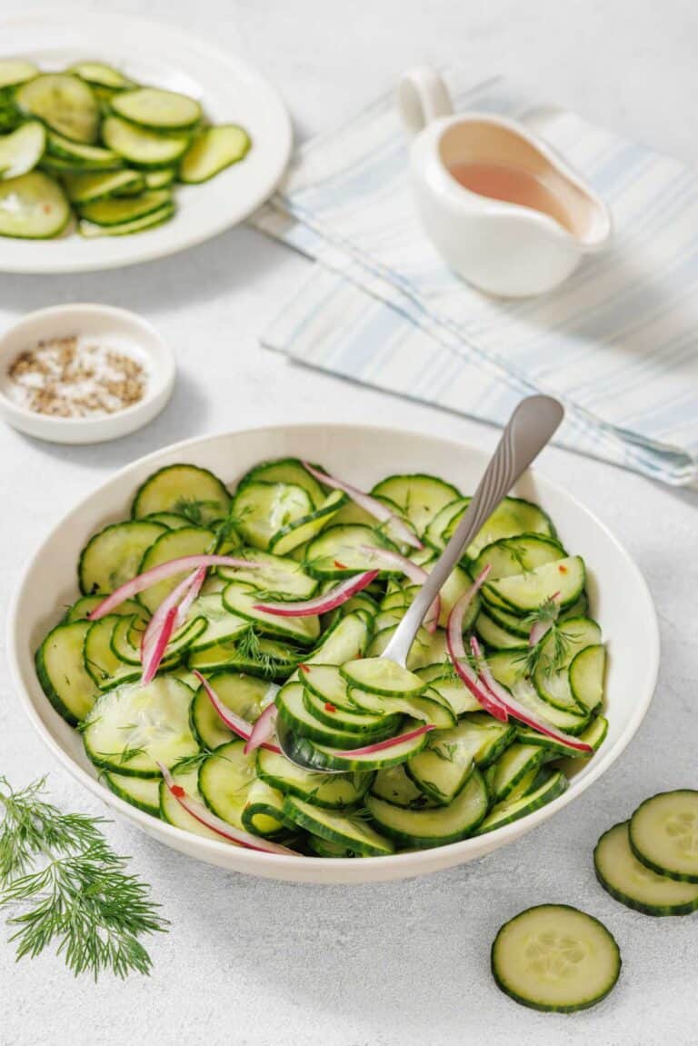 A bowl of Quick Marinated Cucumbers with red onion and dill, a serving spoon, and plates with sliced cucumbers on a light surface. A small dish of salt and dressing are also nearby.