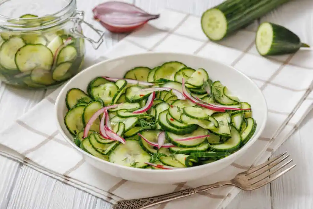 A bowl of Quick Marinated Cucumbers with sliced red onions and herbs, placed on a white cloth with a fork nearby; extra salad and cucumbers are in the background.
