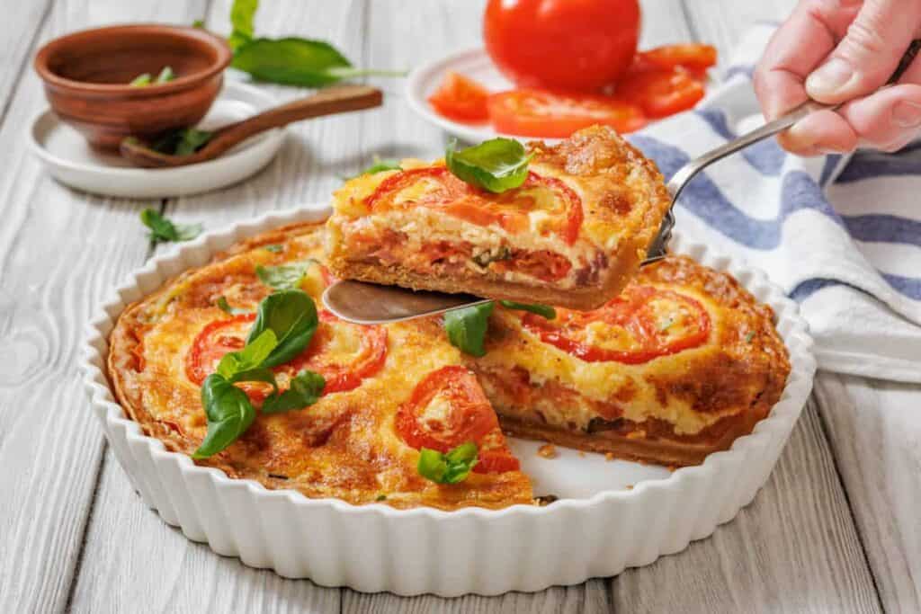A slice of tomato pie being lifted with a serving utensil from a white fluted dish. A whole tomato pie is visible in the dish, garnished with tomato slices and basil.