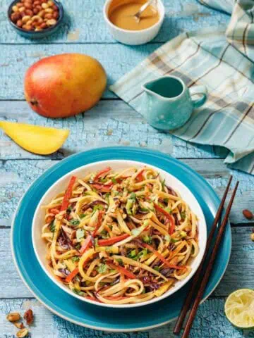 A bowl of colorful vegetable noodles with chopped peanuts, surrounded by a mango, a bowl of sauce, chopped peanuts, a lemon wedge, and wooden chopsticks on a blue table.