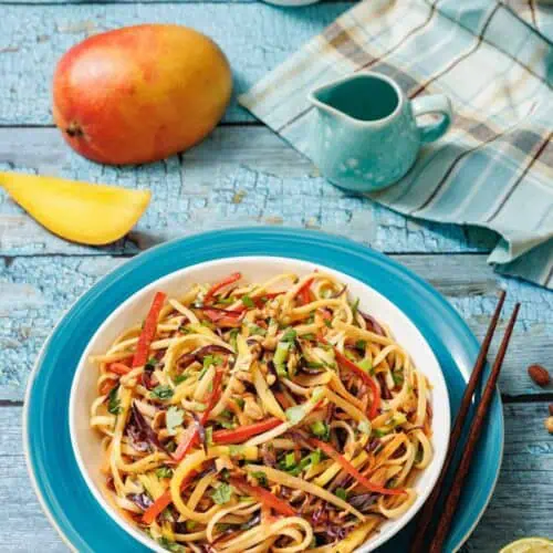 A bowl of colorful vegetable noodles with chopped peanuts, surrounded by a mango, a bowl of sauce, chopped peanuts, a lemon wedge, and wooden chopsticks on a blue table.