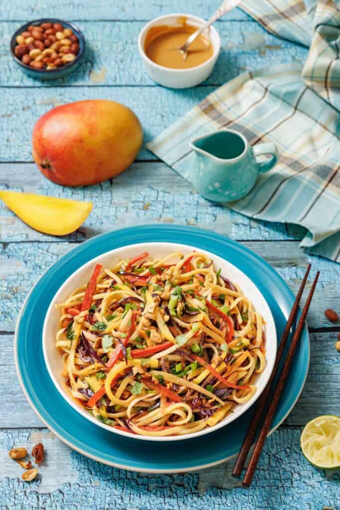 A bowl of colorful vegetable noodles with chopped peanuts, surrounded by a mango, a bowl of sauce, chopped peanuts, a lemon wedge, and wooden chopsticks on a blue table.