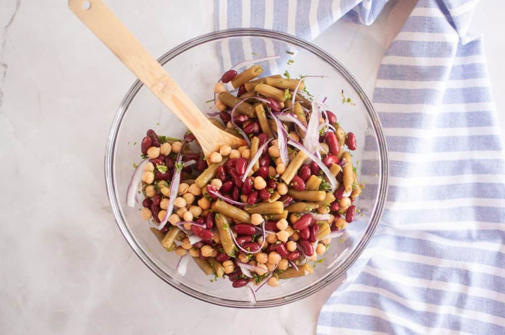 A glass bowl filled with a Three Bean Salad including kidney beans, chickpeas, green beans, and sliced red onion, with a wooden spoon and a striped cloth nearby.