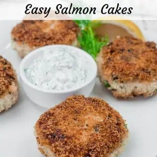 Four Salmon Cakes with a side of creamy dill sauce and a lemon wedge on a white plate.
