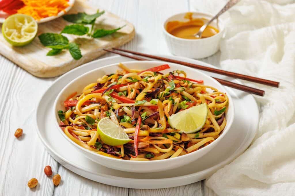 A bowl of stir-fried noodles with vegetables, garnished with lime wedges, served with chopsticks and a side of sauce on a white table.