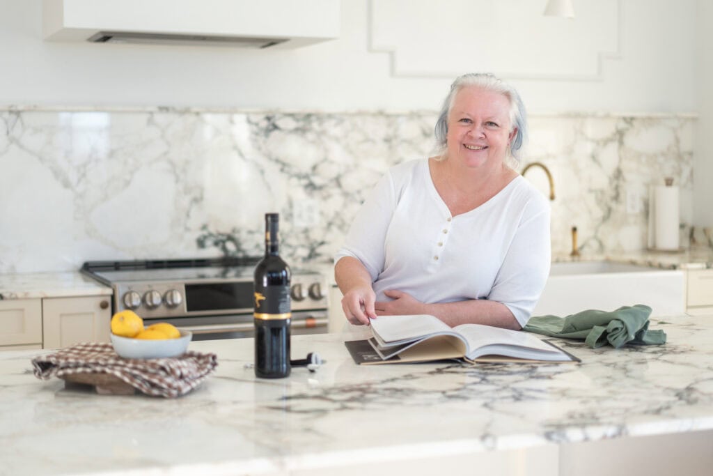 Chef Jenn Allen sits at a marble kitchen counter, smiling, with an open cookbook, wine bottle, lemons, and folded linens in front of her—ready to cook what you love.