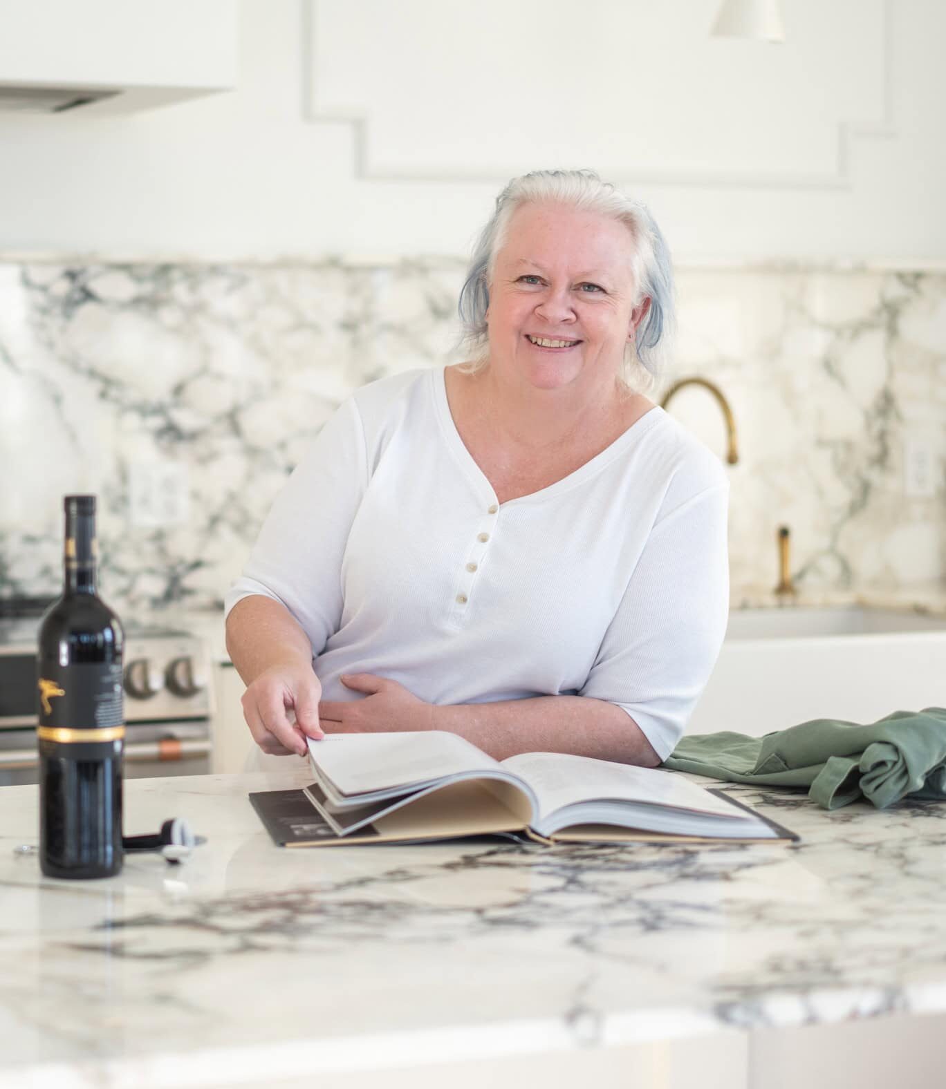 Chef Jenn Allen sits at a marble kitchen counter, smiling, with an open cookbook, wine bottle, lemons, and folded linens in front of her—ready to cook what you love.
