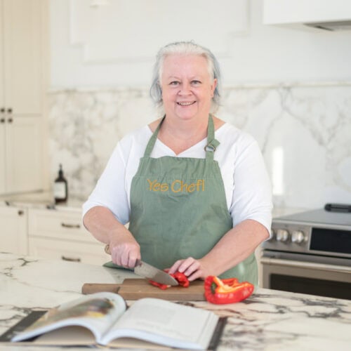 Jenn Allen chops a red bell pepper on a kitchen counter, an open cookbook nearby as she prepares to cook what you love.