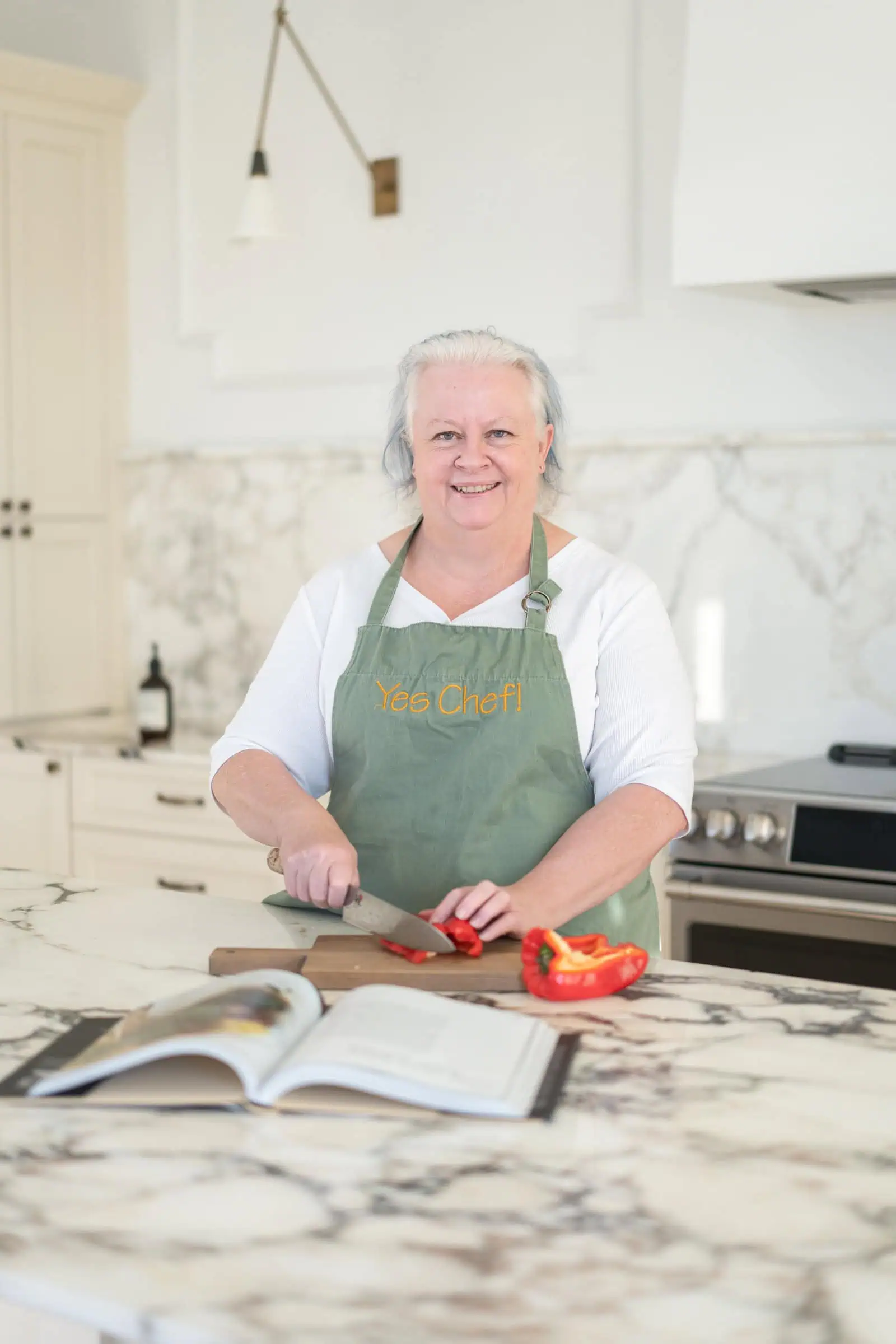 Jenn Allen chops a red bell pepper on a kitchen counter, an open cookbook nearby as she prepares to cook what you love.