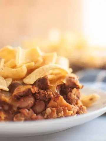 A close up take of Frito Pie in a small white plate on a white table with corn chips on top.