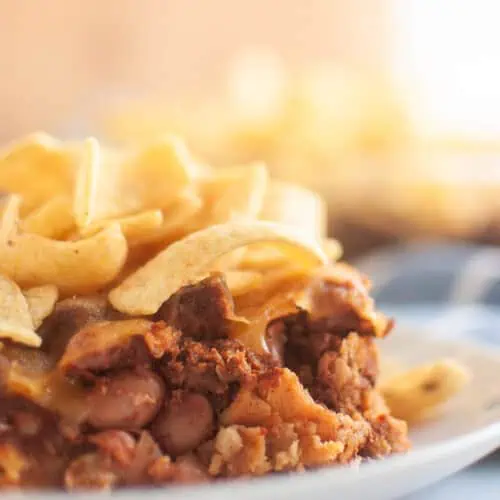 A close up take of Frito Pie in a small white plate on a white table with corn chips on top.