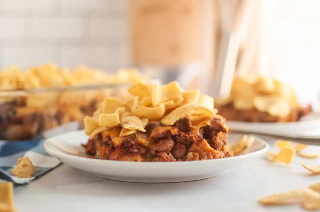 A front take of Frito Pie in a small white plate on a white table with corn chips on top and scattered.