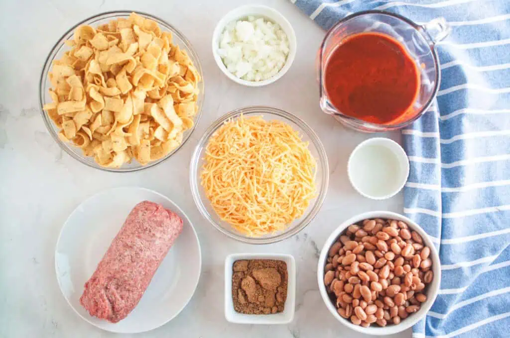 Overhead shot of ingredients for Frito pie, including Fritos, onions, enchilada sauce, cheese, ground beef, spices, oil, and beans, arranged on a light surface with a blue striped cloth.