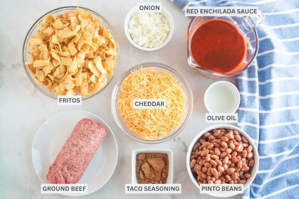 Overhead shot of ingredients for Frito pie, including Fritos, onions, enchilada sauce, cheese, ground beef, spices, oil, and beans, arranged on a light surface with a blue striped cloth and labels for each ingredient.