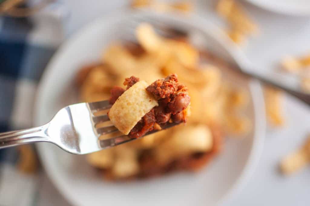 A close up take of Frito Pie in a fork with a blurry take of a small serve in a white plate in the background.