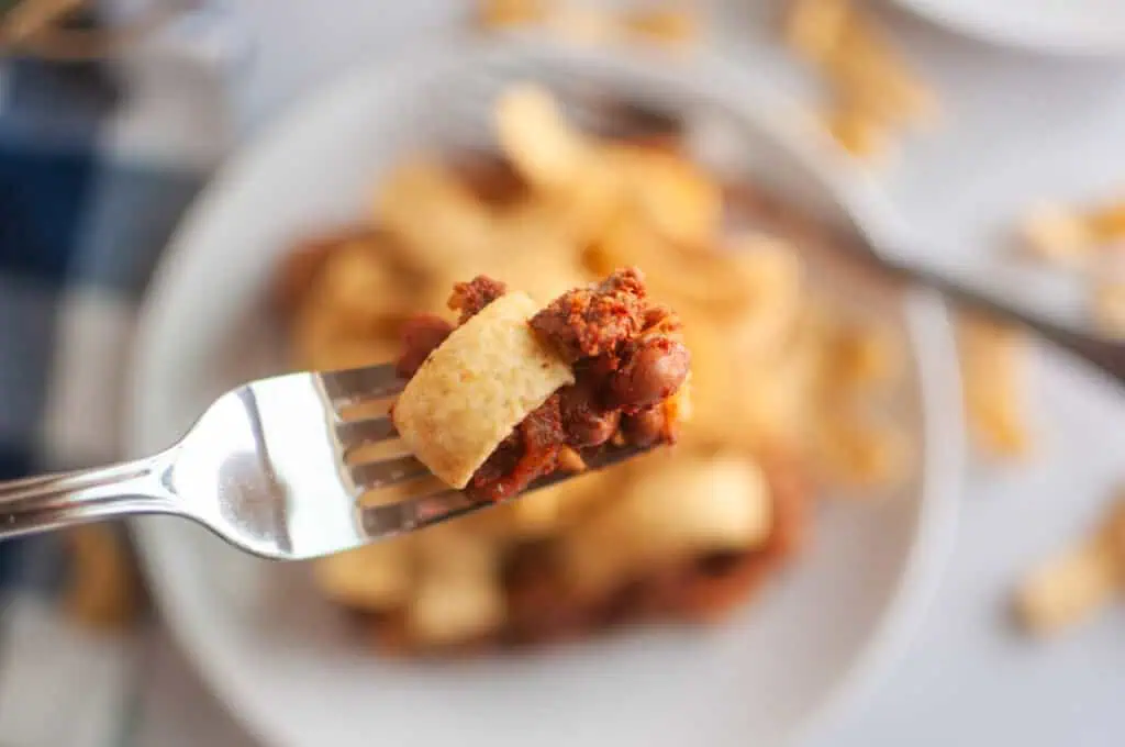 A close up take of Frito Pie in a fork with a blurry take of a small serve in a white plate in the background.