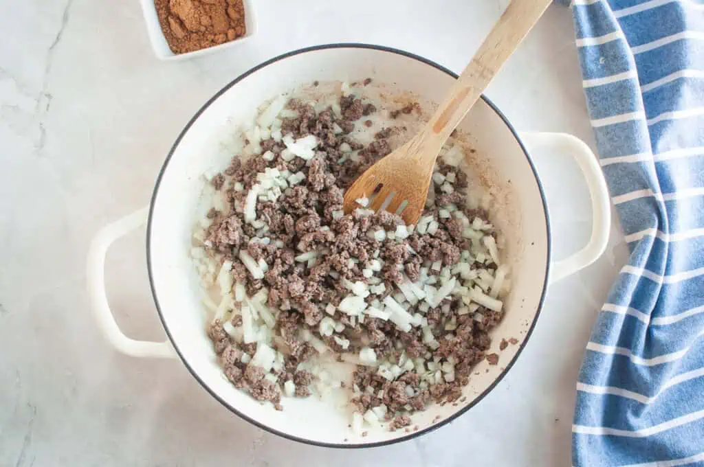 Overhead shot of cooked ground beef and chopped onions in a white pot with a wooden spoon, with a bowl of spices nearby.