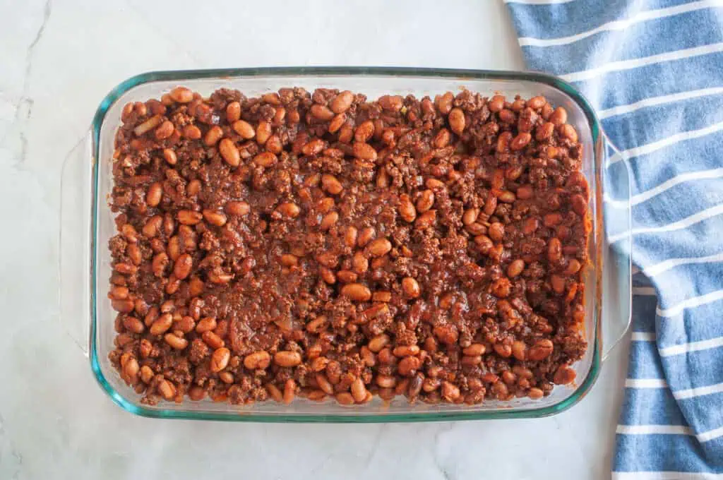 Overhead shot of baked beans and ground beef mixture in a clear glass baking dish, with a blue striped cloth beside it.